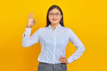 Portrait of beautiful and smilling young business woman with confident face making okay sign with finger on yellow background