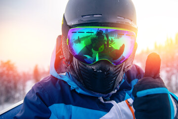 Skier man in balaclava, helmet and glasses is taking selfie in snow on mountain ski lift, sun light