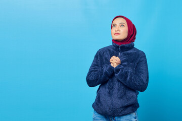 Attractive young Asian woman praying hopefully and looking up on blue background