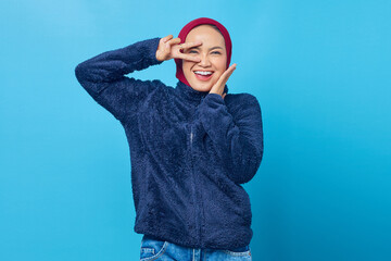 Cheerful young Asian woman showing peace sign over eyes and palms touching cheek on blue background
