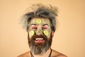 Funny man closeup with clay mask and cucumber slices on face. Spa, dermatology, wellness and facial treatment concept. Man having cosmetic moisturizing mask isolated studio background. Male face care.