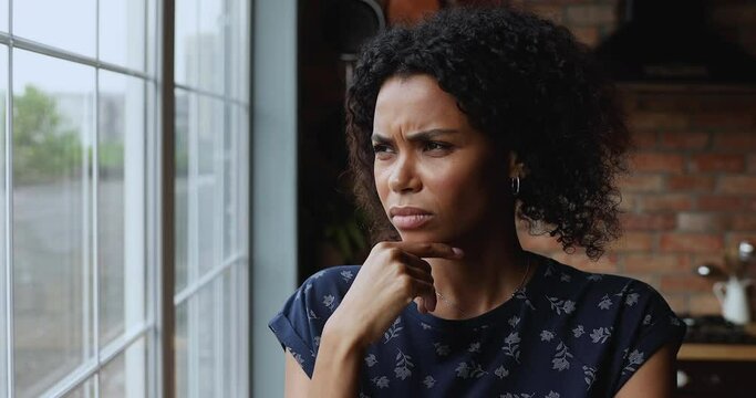 Close up African woman feels troubled due personal life problem, is nervous touch chin thinks over concerns, standing alone in kitchen looks out window. Negative emotions, heartache, jealousy concept
