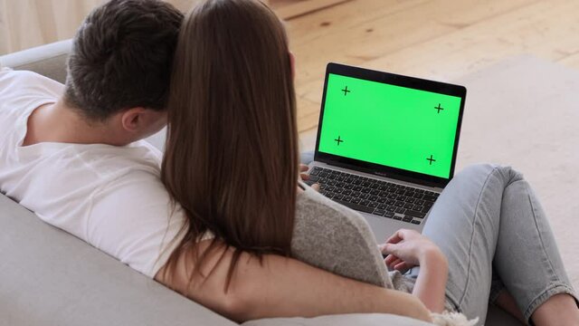 Young couple in love using laptop and hugging while sitting on couch in apartment interior spbi. Back view of beautiful woman, man look at computer screen and hug, watch web or video and sit on sofa