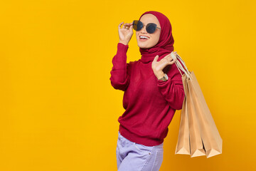 Cheerful young Asian woman wearing sunglasses and holding shopping bags on yellow background