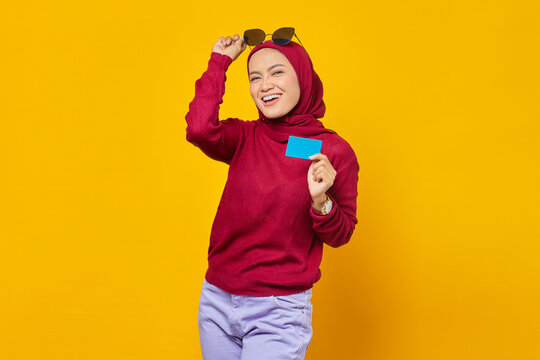 Smiling young Asian woman holding credit card and take off spectacles on yellow background
