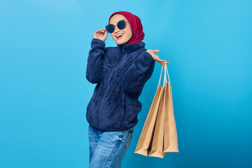 Cheerful young Asian woman wearing sunglasses and holding shopping bags on blue background