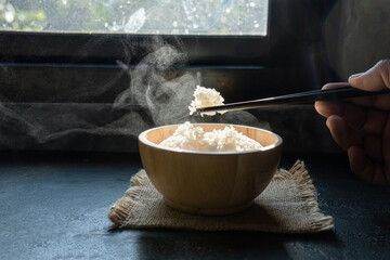 The man hand of using black chopsticks holding hot jasmine rice with smoke and steam in black bowl over dark background.hot food concept