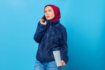 Pensive young Asian woman talking on mobile and holding laptop on blue background