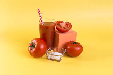 Natural tomato juice in a transparent glass with a decor of tomatoes, salt and a colored cube on a yellow background close-up