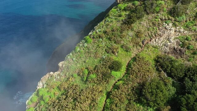 Drone shot of a young, fit and strong man hiking up Espigao Amorelo on Maderia during the summer. Thin clouds are drifting by with the blue and turquoise ocean below in the background of the cliff.