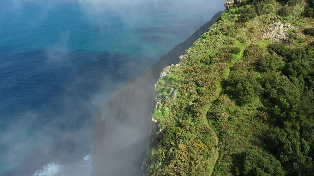 Drone shot going upwards of a young, fit and strong man hiking up Espigao Amorelo on Maderia during the summer. Thin clouds are drifting by with the blue and turquoise ocean below in the background.