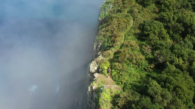 Drone shot of a young, fit and strong man hiking up Espigao Amorelo on Maderia during the summer. Walking right next to the steep edge with a mysterious fog and mist around him.