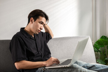 Tired Young man student or businessman thinking a solution for work problems working on a computer laptop at a home office for video conference, sits on a couch, looking financial crisis on a screen