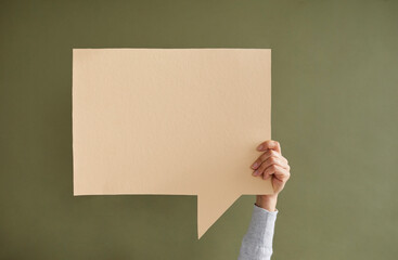 Young anonymous woman holding one empty beige square speech bubble in hand on green background. Blank paper card layout with copy space for message, quote, inspiration, opinion or vote.