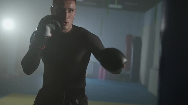 Aggressive fighter trains his punches and defense in the boxing gym, a man boxer beats a punching bag, 4k slow motion.