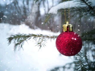 A red Christmas ball is hanging on the Christmas tree. It's snowing in the winter forest. Christmas card.