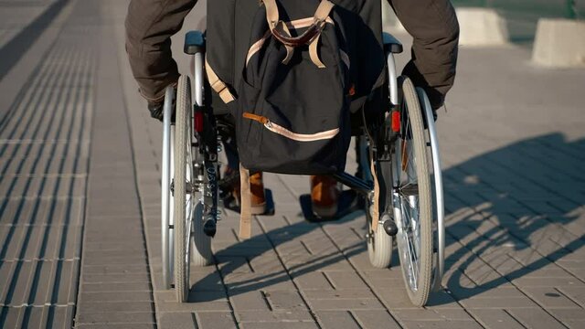 man in wheelchair is moving over street at sunny weather, rear view