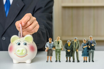 Saving for retirement and pension fund concept : Senior retired couple stand near a piggybank, a man insert money in a deposit box for future expenses, depicting long-term investment for aging society - obrazy, fototapety, plakaty