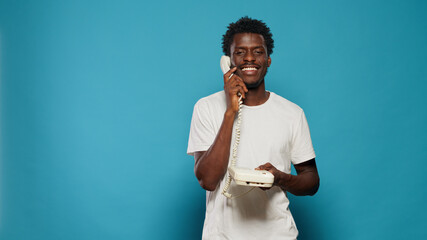 Young adult using landline telephone for phone call. Man talking on retro phone and smiling, using landline phone for communication. Modern person having fun and speaking on call - obrazy, fototapety, plakaty