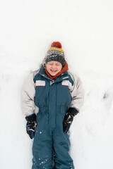 Grimacing boy in a warm jumpsuit and hat lies in the snow. Winter holidays. Child is playing in the snow. Top view