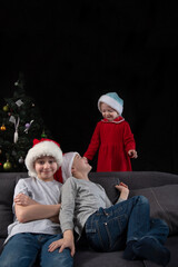 Fototapeta premium Siblings at home on the sofa on Christmas Eve. Happy children play on the sofa near Christmas tree. Vertical frame
