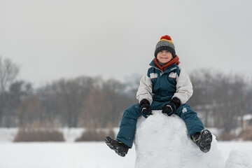 Fototapeta na wymiar Smiling boy of seven years old sits on snow tower. Child builds snow castle. Winter time.