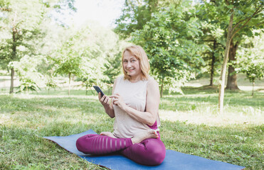 an elderly yogi woman is sitting in a lotus position in nature with a phone in her hands