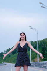 A happy girl with long hair in a black dress is standing on the road, hands out to the sides