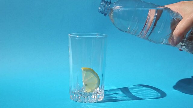 A close-up of a glass with a slice of lemon on a bright blue background, into which clean drinking water is poured from a plastic bottle. Healthy food, healthy drinks, the wellness concept
