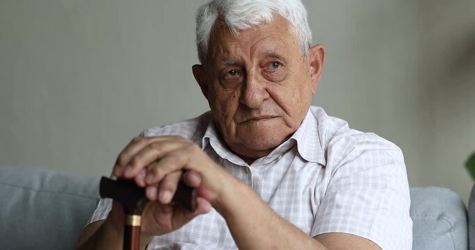 Serious sad senior age male patient who use walking cane after orthopedic trauma sit on sofa wait for doctor appointment need help care rehabilitation. Old man having disability lost in heavy thoughts