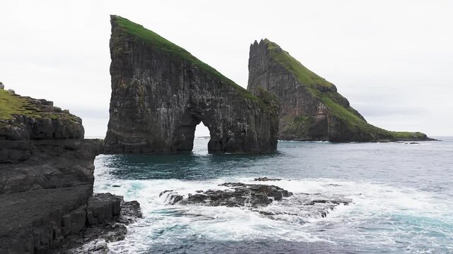 Aerial view of the Rocky Drangarnir sea stack in the Faroe Islands and the skarosafossur Waterfall.Beautiful pristine nature, rough sea, rock cliffs,waves create sea foam, clouds in sky 