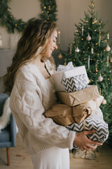 Young beautiful woman holding gift boxes and presents in a cozy room with Christmas tree on background. Holiday preparations.