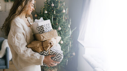 Young beautiful woman holding gift boxes and presents in a cozy room with Christmas tree on background. Holiday preparations.