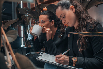 Muchacha joven de oscuro concentrada en sus contratos y escribiendo planteamientos e ideas junto a su compañera de trabajo mientras beben café en una cafeteria 