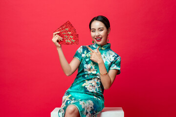 Portrait of smiling happy Asian woman in oriental cheongsam costume pointing to red envelopes or...