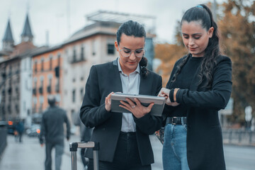 Mujeres secretarias jóvenes de cabello oscuro formales con una sonrisa de pie firmando papeles con contratos junto su compañera en el centro de la ciudad mientras mira la hora para llegar a su trabajo