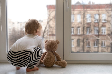 Cute caucasian blonde baby girl about 1,2 years old with teddy bear sitting on sill looking through window at street autumn view.Infant,toddler,kid safety,secuity.Lock down,isolation,stay home concept