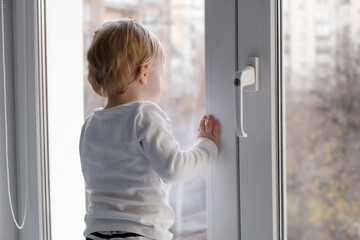 Fototapeta na wymiar Cute caucasian blonde baby girl about 1,2 years old on sill looking through window with handle lock at street autumn view.Infant,toddler,kid safety,secuity..Lock down,isolation,stay home concept