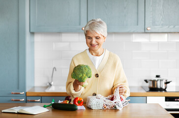 healthy eating, food cooking and culinary concept - happy smiling woman with vegetables in string bag and cook book on kitchen