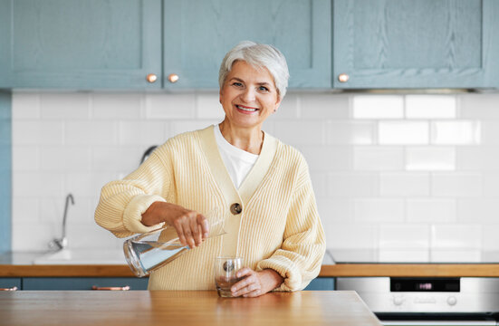 Leisure And People Concept - Happy Smiling Woman Pouring Water From Jug To Drinking Glass On Kitchen At Home