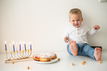 Kid in white t-shirt on white background celebrating Hanukkah. Jewish festival of lights. Child smile and happy. on table donuts and traditional menorah with lighting candles. holiday in Israel 
