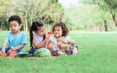 Obraz premium Three mixed race caucasian little cute children kids sitting, playing in outdoor green park for picnic, eating fruit, watermelon and pineapple with freshness. Education, Friendship, Diversity Concept.