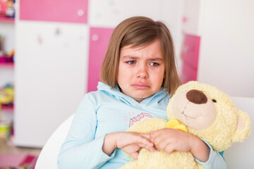 Cute little caucasian crying girl with teddy bear in bedroom. Sad unhappy children emotions