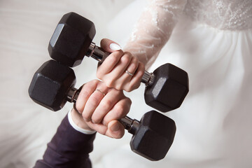 the hands of the newlyweds with wedding rings hold dumbbells