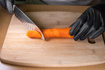 Chef's hands in black gloves holding a knife and cutting peeled carrots on a kitchen wooden board. The process of preparing