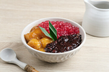 Indonesian traditional dessert. Black glutinous rice with Candil and Sagu (Mutiara) porridge and pandan leaf. Served in a white bowl on wooden table. Blurred background and selective focus.
