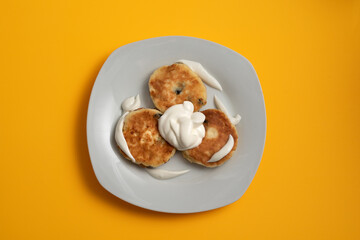 Three cottage cheese pancakes (syrniki) on white plate on yellow background with sour cream poured over