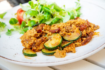 Vegan breakfast prepared with scrambled tofu and eggs, cooked with dried tomatoes and zucchini served with fresh lettuce and cucumber in white ceramic vintage plate