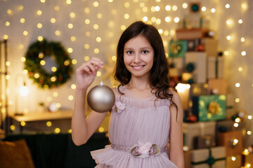 Fototapeta premium cute girl in festive dress holding christmas decoration in decorated room with heap of christmas gifts