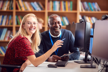 Female University Or College Student Working At Computer In Library Being Helped By Tutor